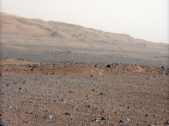 Foto feita pelo jipe Curiosity em Marte mostra solo pedregoso perto do local da aterrissagem e, mais adiante, a base rochosa do monte Sharp