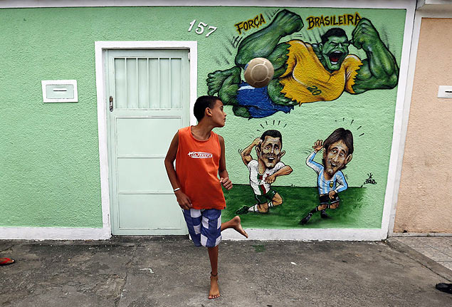 Menino joga bola diante de muro pintado com referncia aos jogadores Hulk, Cristiano Ronaldo e Messi em rua do Rio