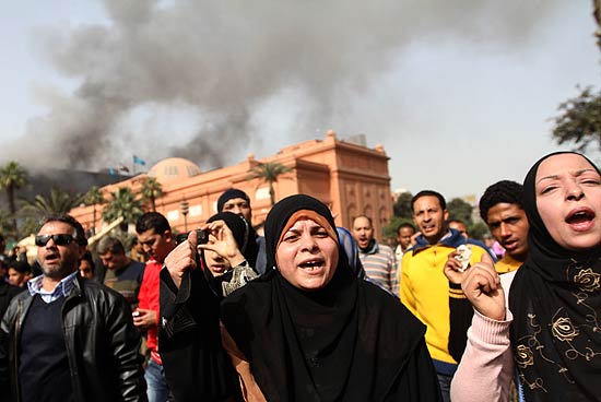 Mulheres participam dos protestos no Cairo; ao fundo, sede do partido do ditador Hosni Mubarak  incendiada