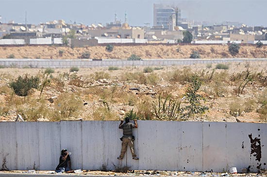 Rebeldes olham por cima de muro para acompanhar a batalha pelo complexo de Bab Al-Aziziya, em Trípoli