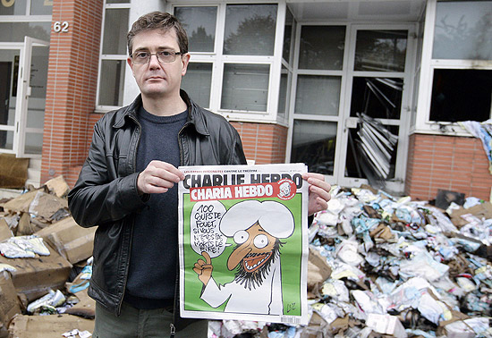 Editor da "Charlie Hebdo" mostra capa com charge de Maom; ao fundo, redao da revista que foi bombardeada