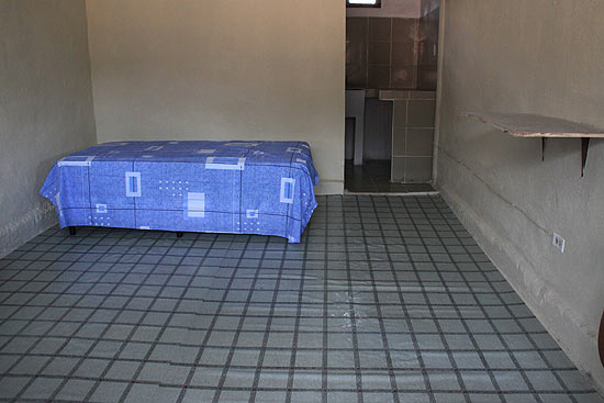 Cela de 12 metros quadrados, com duas janelas, uma porta de metal, uma cama de solteiro e uma latrina