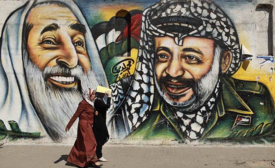 Mulheres passam em frente a muro com retrato do lder palestino Yasser Arafat ( direita) na Faixa de Gaza