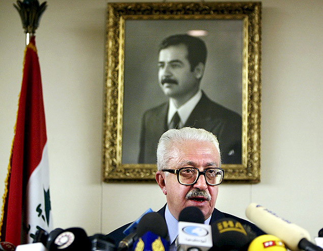 Tariq Aziz d entrevista com a foto de Saddam Hussein ao fundo 