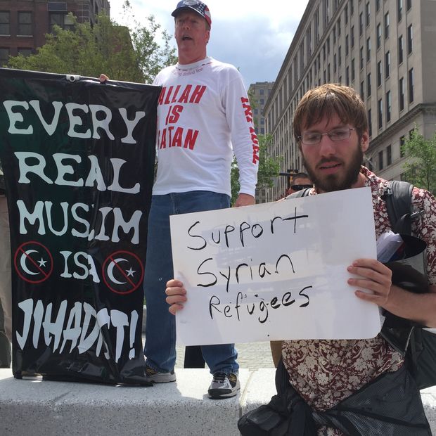 Homem segura cartaz de apoio a refugiados srios; ao fundo, manifestante anti-isl 
