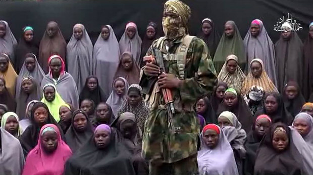 TOPSHOT - This video grab image created on August 14, 2016 taken from a video released on youtube purportedly by Islamist group Boko Haram showing what is claimed to be one of the groups fighters at an undisclosed location standing in front of girls allegedly kidnapped from Chibok in April 2014. Boko Haram on August 14, 2016 released a video of the girls allegedly kidnapped from Chibok in April 2014, showing some who are still alive and claiming others died in air strikes. The video is the latest release from embattled Boko Haram leader Abubakar Shekau, who earlier this month denied claims that he had been replaced as the leader of the jihadist group. / AFP PHOTO / HO / RESTRICTED TO EDITORIAL USE - MANDATORY CREDIT 