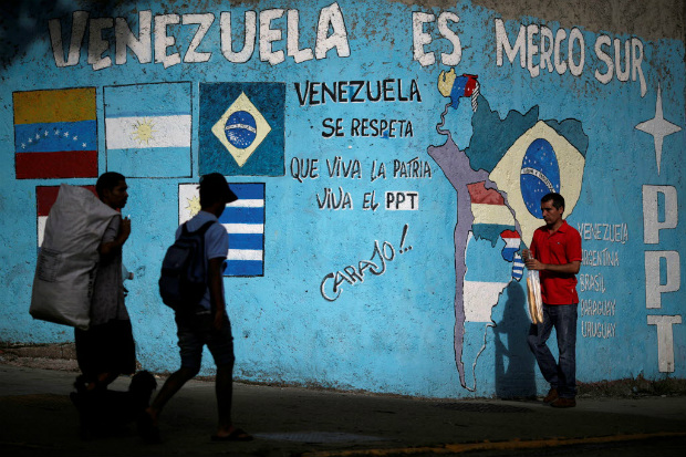 Pessoas passam em frente a um muro com o dizer 'Venezuela  Mercosul', em Caracas