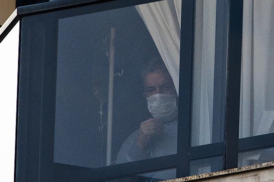 O ex-presidente Lula aparece na janela do seu apartamento em Sao Bernardo do Campo, com mascara cirurgica e, ao fundo, um uporte para medicamento