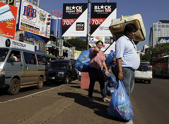 Pessoas carregando compras em Ciudad del Este; ao fundo, anncio da Black Friday, que acontecer em maio