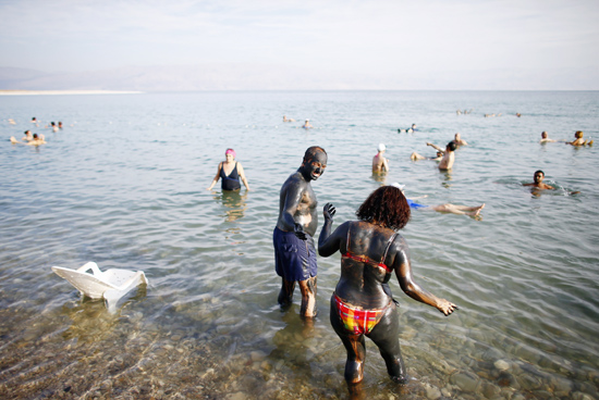 Casal coberto de lama negra entra no Mar Morto, enquanto outros visitantes boiam na gua 