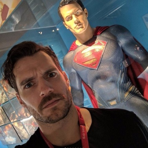 Henry Cavill, de bigode, posa em frente ao seu traje de Superman 