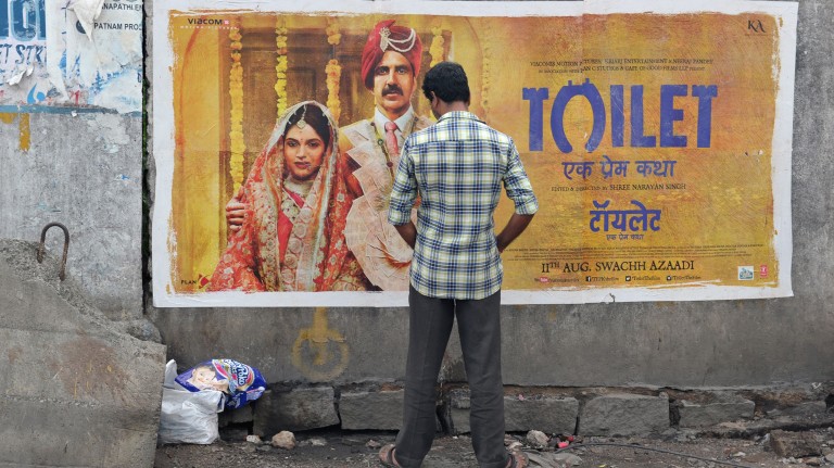 Indiano urina em parede na beira da estrada em frente a cartaz do filme "Toilet" 