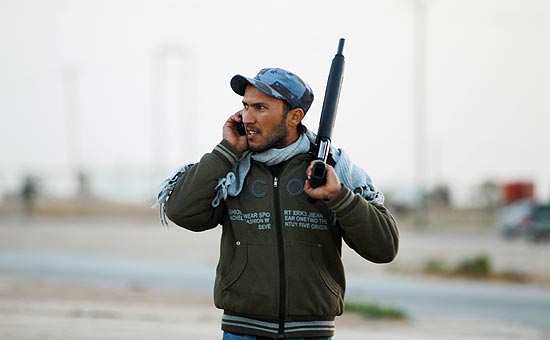Policial líbio fala ai telefone enquanto vigia bloqueio em estrada rumo ao leste, dominado pela oposição