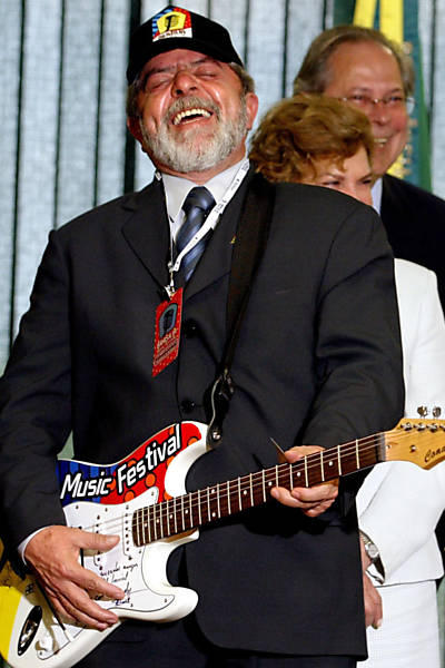 O presidente Luiz Inácio Lula da Silva finge tocar guitarra, com a primeira-dama Marisa Letícia Lula da Silva, e o ministro-chefe da Casa Civil, José Dirceu, ao fundo, em Brasília (DF); a guitarra será leiloada, e o dinheiro obtido com a venda será doado para o programa Fome Zero. Lula ganhou a guitarra autografada e um boné do festival Brasília Music Festival durante encontro com os integrantes da banda Capital Inicial