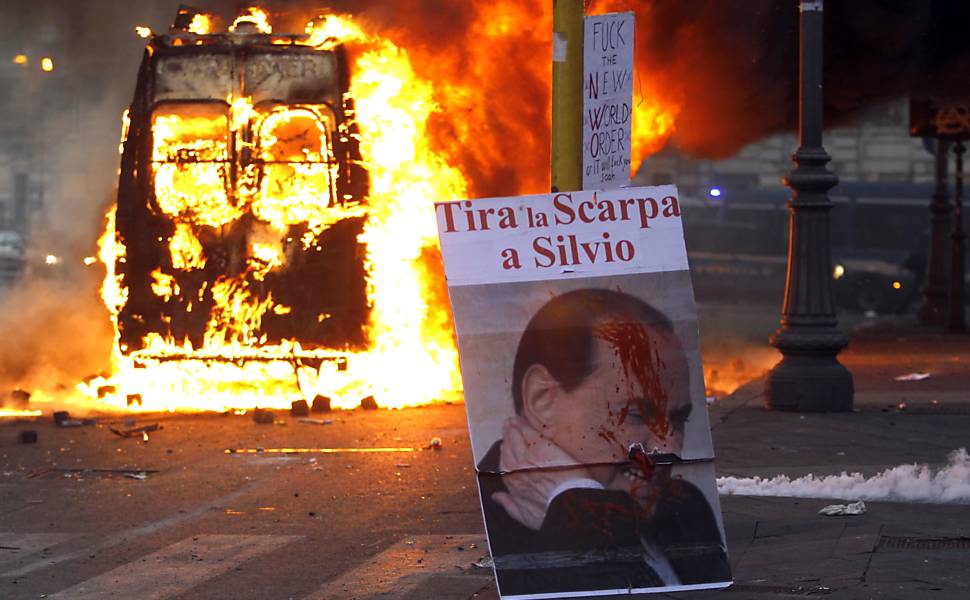 Cartaz com foto do premiê italiano, Silvio Berlusconi, é visto enquanto viatura da polícia queima em chamas durante ato do grupo Indignati (indignados), em Roma