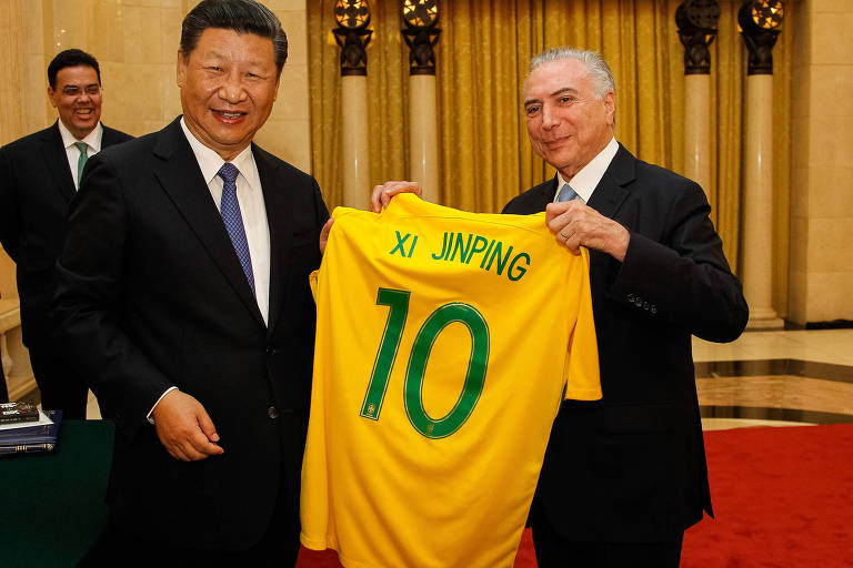 Temer segura a camisa amarela da Seleção Brasileira, com o número 10 e o nome de Xi Jinping ao lado do líder chinês. Ambos sorriem. Os dois usam terno preto e camisa branca. Ao fundo, a parede amarelada do Grande Salão do Povo, que tem tapete vermelho. À esquerda aparecem dois seguranças de terno.