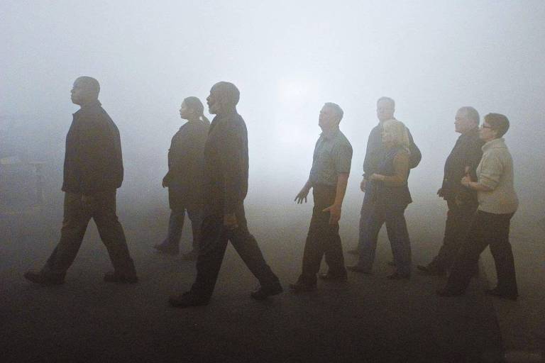 Silhuetas de pessoas emergem de uma névoa densa. A visibilidade limitada e a disposição das figuras sugerem uma caminhada coletiva através de um espaço indefinido