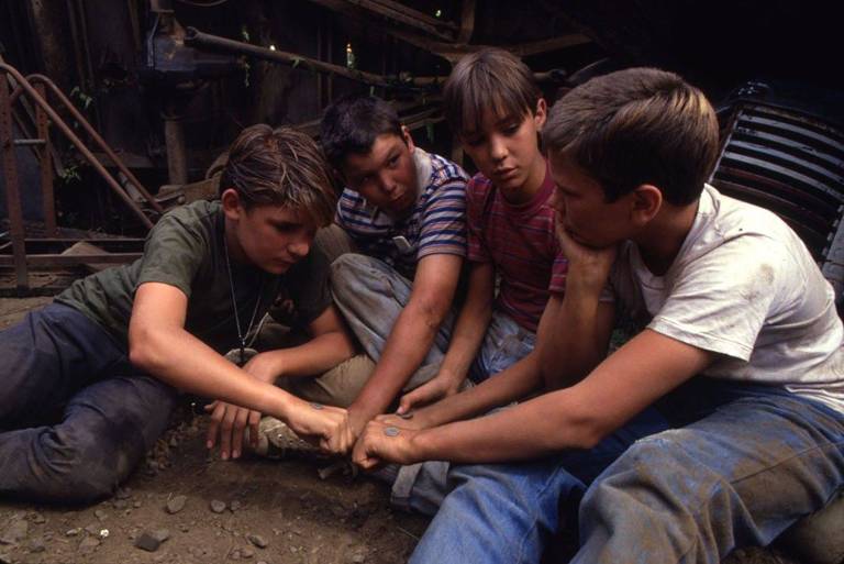 Quatro garotos, vestindo calças jeans e camisetas, estão sentados em um chão de terra. Eles estão com as mãos juntas, ao centro, com os punhos cerrados