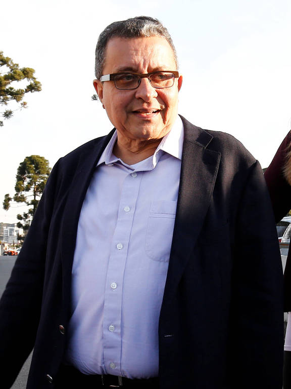 O marqueteiro João Santana aparece em primeiro plano, vestindo uma camisa azul clara por baixo de um blazer preto. Santana está olhando para seu lado esquerdo, com um leve sorriso no rosto.