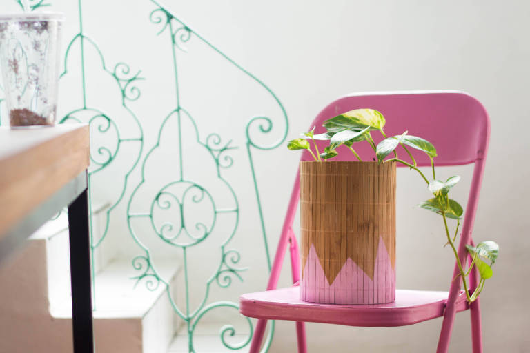 Vaso de planta de palha com desenho geométrico rosa em sua base. Está em cima de uma cadeira metálica rosa em frente a usa escada
