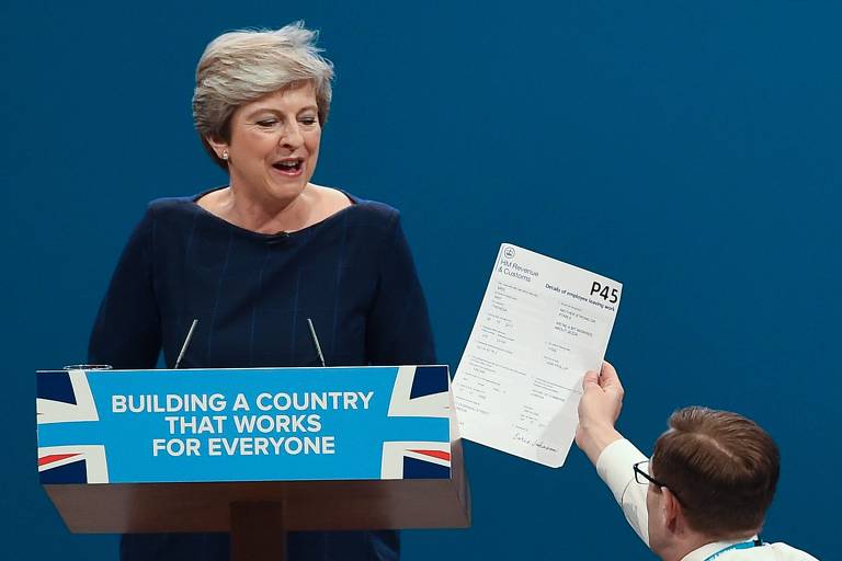 Protester comedian Simon Brodkin (R) gives a piece of paper written as a mock P45 (employee leaving form) to Britain's Prime Minister Theresa May (L) as she was delivering her speech on the final day of the Conservative Party annual conference at the Manchester Central Convention Centre in Manchester, northwest England, on October 4, 2017. The protester interrupted the leader's speech to hand her a paper and then was escorted out of the auditorium. / AFP PHOTO / PAUL ELLIS