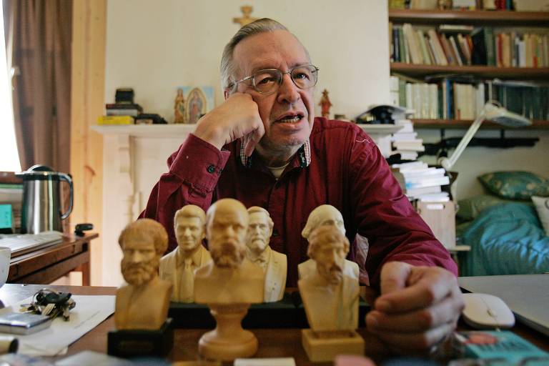 Olavo exibe bustos em miniaturas de filósofos, está sentado à mesa, ao fundo estantes com livros