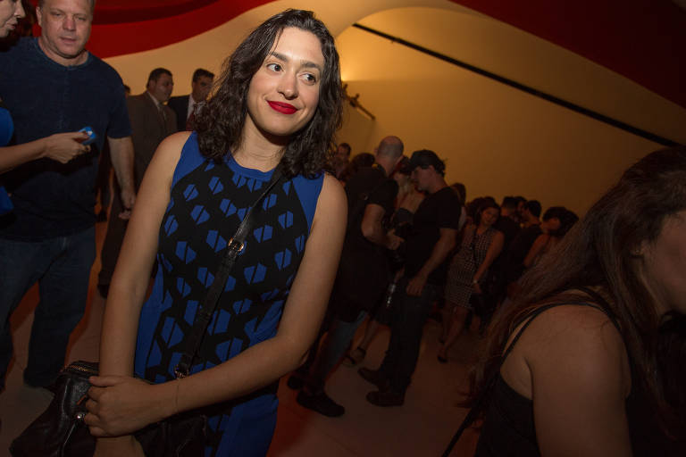 Moça de vestido azul estampado e boca com batom vermelho-vivo sorri sem olhar direto para a câmera; ao fundo, um grupo de pessoas no hall de uma sala de cinema
