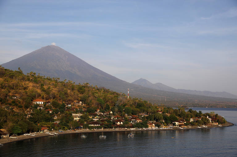 A imagem mostra uma foto aérea de Bali, na Indonésia. Ao fundo e à distância está o vulcão Agung. Atrás dele, é possível ver dois outras montanhas mais distantes. À frente, um monte com árvores e pequenas casas, que acabam na praia