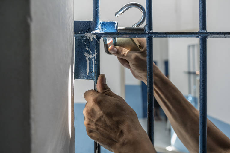 Duas mãos abrem um cadeado no portão de uma cela 