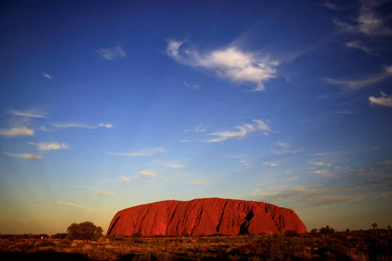 O monólito Uluru é visto a distância. Ele tem encostas altas, como morros, mas o topo é plano, parecendo uma mesa. A cor dele é um vermelho bem vivo, semelhante ao terreno arenoso em que se encontra, no meio do deserto australiano