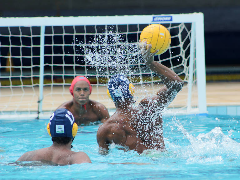 Atletas jogam polo aquático em piscina. Três esportistas homens aparecem em jogo, sendo que um deles está com o braço esticado prestes a lançar a bola em direção ao gol.