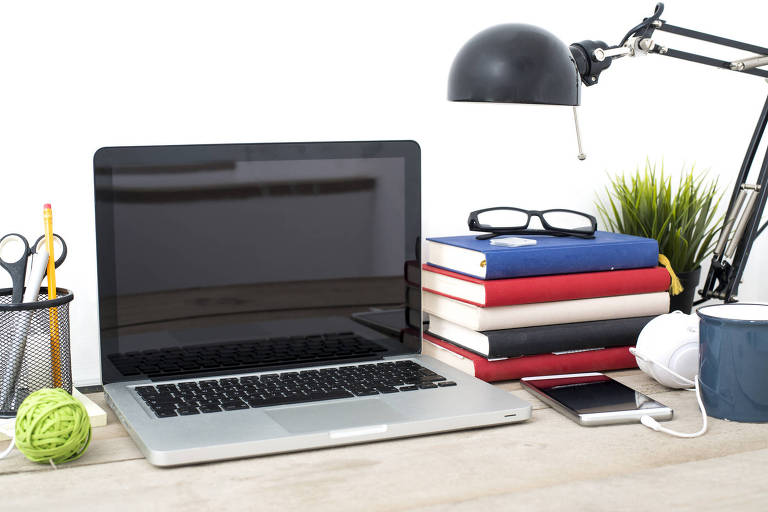Foto mostra um laptop sobre uma mesa, com a tela levantada. H uma pilha de livros ao lado e uma luminria