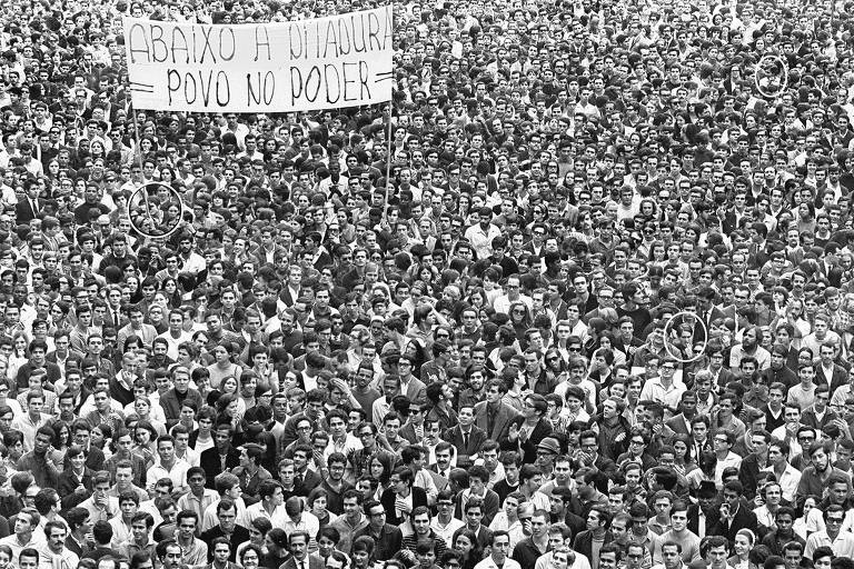 Foto mostra multidão marchando. No canto superior esquerdo, uma faixa diz "Abaixo a ditadura. Povo no poder"