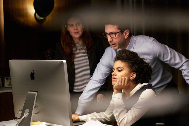 Observadors por uma produtora, o ator  David Schwimmer, de camisa e gravata, se debruça sobre uma mesa com um computador diante da qual está sentada a atriz Zazie Beetz durante filmagem de cena da série "That's Harassment"