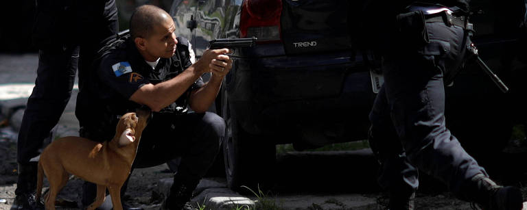 Policial militar em posição de tiro durante tiroteio na Cidade de Deus, zona oeste do Rio, nesta quinta (1); enquanto se posiciona de joelhos, um pequeno cachorro se aproxima do PM