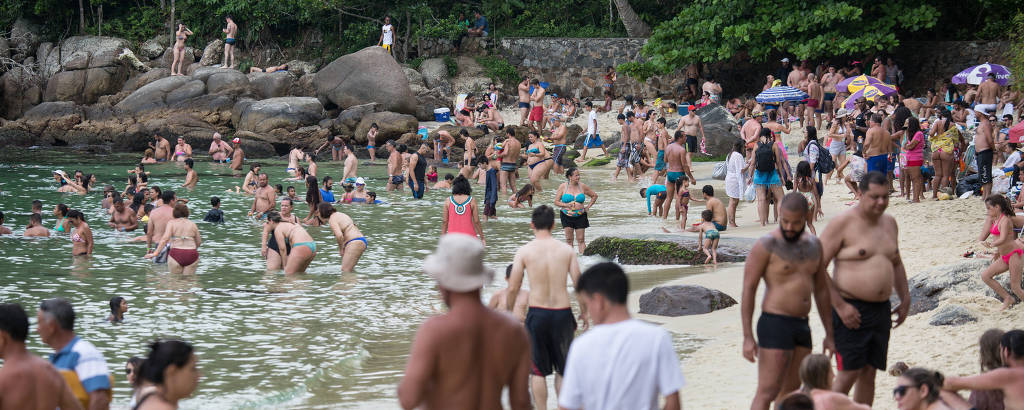 Multidão de turistas sentados na areia, brincando no mar e subindo em pedras em uma praia estreita na Ilha das Couves, em Ubatuba, no litoral norte de SP