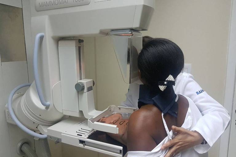 Mulher, vista de costas, coloca mama esquerda em aparelho de mamografia. Ela é auxiliada por pessoa usando jaleco branco.