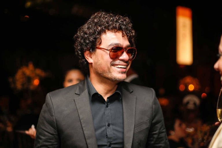 De paletó e camisa social pretos e óculos escuros com armação e lentes vermelhas, cantor Léo Maia posa para foto. Ele sorri enquanto olha para a direita