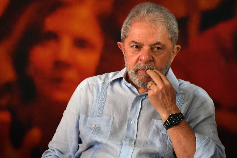 Lula sentado de camisa azul diante de painel vermelho
