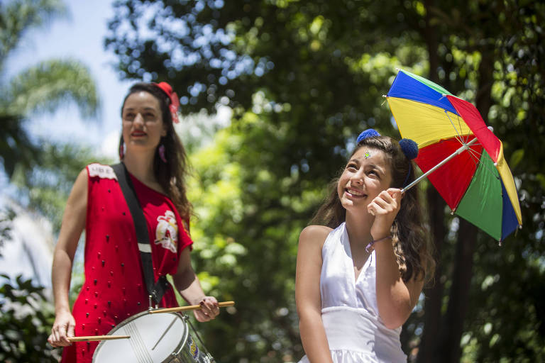 A corretora Thais Haliski com vestido vermelho, leva um tambor, ao lado da filha Valentina, que sorri em um vestido branco e um guarda-chuvas colorido