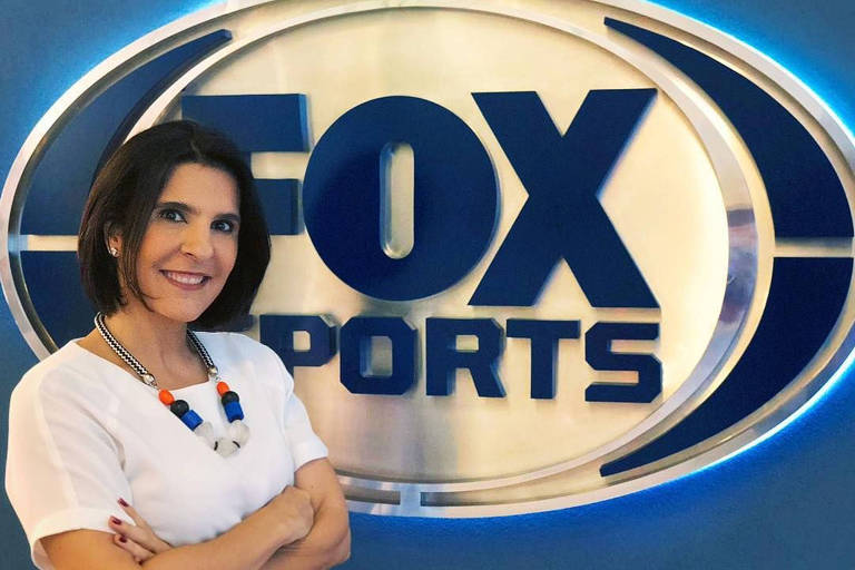 A jornalista Vanessa Riche, de blusa branca e braços cruzados, em frente ao logo do canal Fox Sports