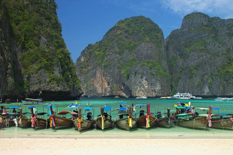 A foto mostra uma fileira com dez barcos compridos típidos da Tailândia atracados na areia da praia. Ao fundo, há pelo menos três montanhas rochosas que formam a baía de Maya Bay