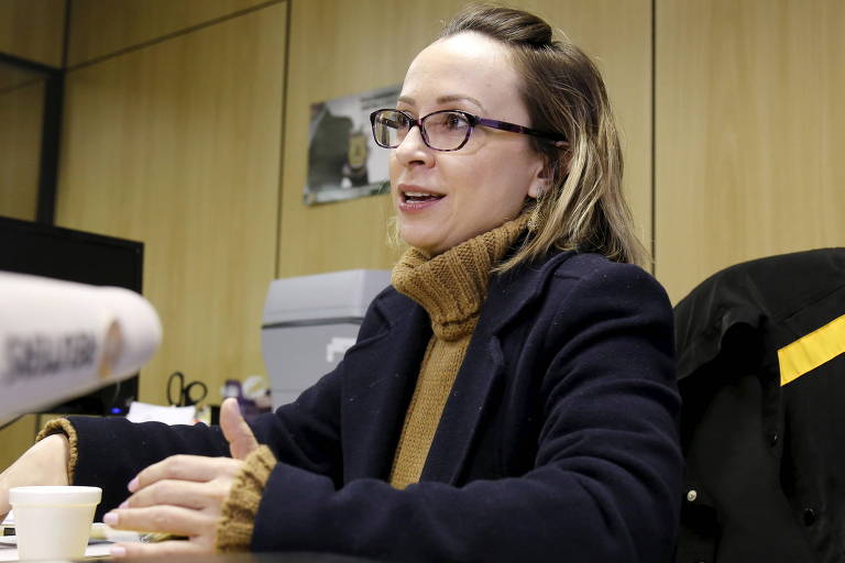 Delegada Erika Marena sentada a sua mesa no gabinete da PF. Ela é loira e usa óculos. Em sua cadeira, um colete com o distintivo da PF está pendurado. 