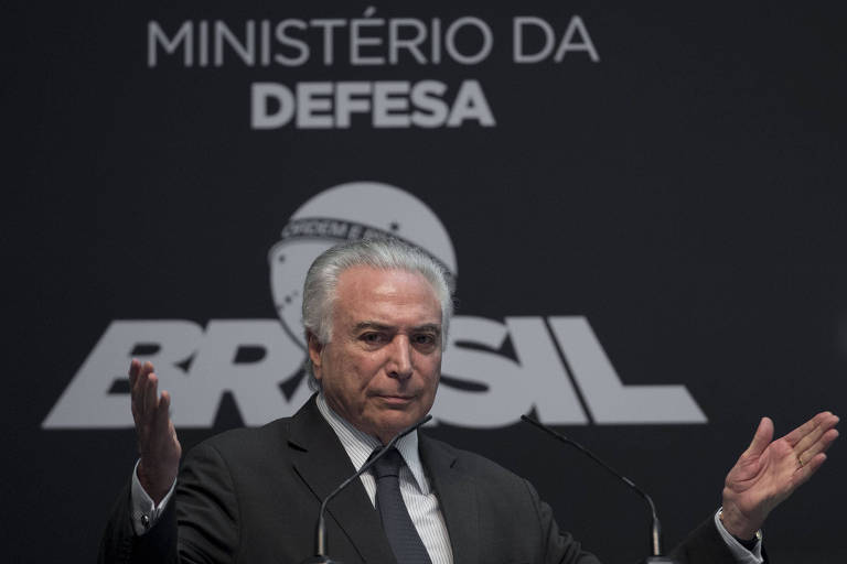 O presidente Michel Temer abre os braços durante evento no Rio de Janeiro; atrás, um banner com o escrito "Ministério da Defesa"