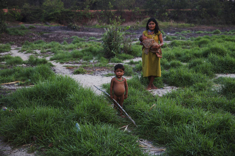 Mulher indígena carrega criança no colo em meio a pastagem, logo atrás de criança nua segurando uma lança