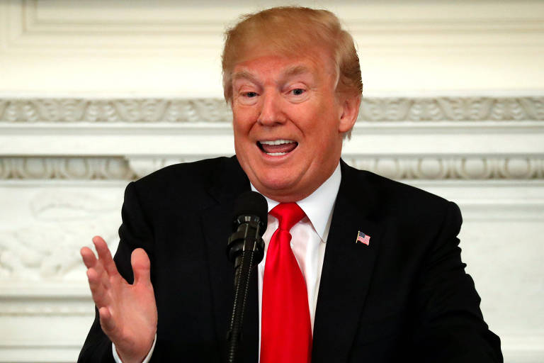 Sentado diante de uma parede, Donald Trump, de gravata vermelha e terno preto, gesticula com a mão direita enquanto fala durante reunião na Casa Branca