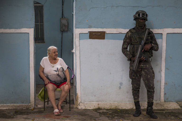 Soldado armado ao lado de senhora sentada na porta de casa