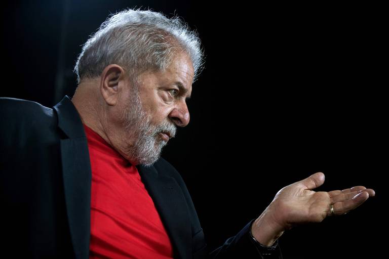 O ex-presidente Luiz Inácio Lula da Silva fotografado de perfil direito, do peito para cima, usando blazer preto e camiseta vermelha, fala e estende a mão esquerda para o interlocutor que não aparece no quadro