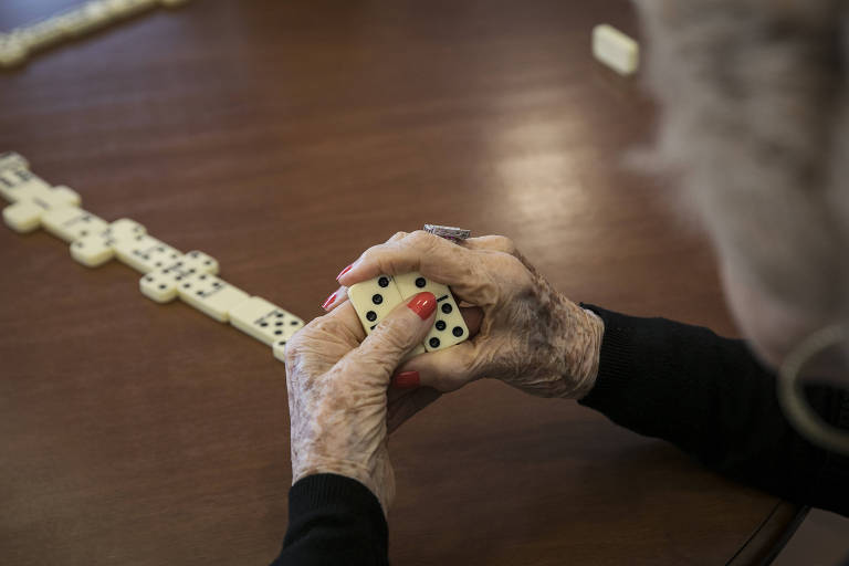 Imagem destaca as mãos de uma mulher idosa, com as unhas pintadas de vermelho e segurando peças de dominó