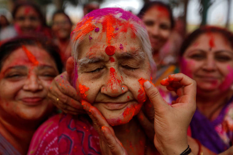 Na Índia, mulher sorridente fecha os olhos enquanto pessoas passam pó colorido em seu rosto